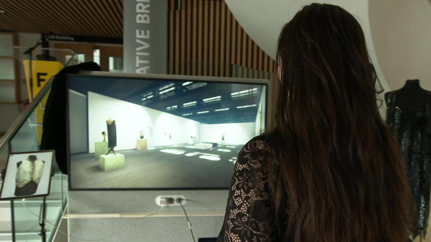 A woman can be seen from behind. She has long, dark hair and is wearing a black lace top. She is standing in front of a screen on which part of the digital exhibition can be seen.