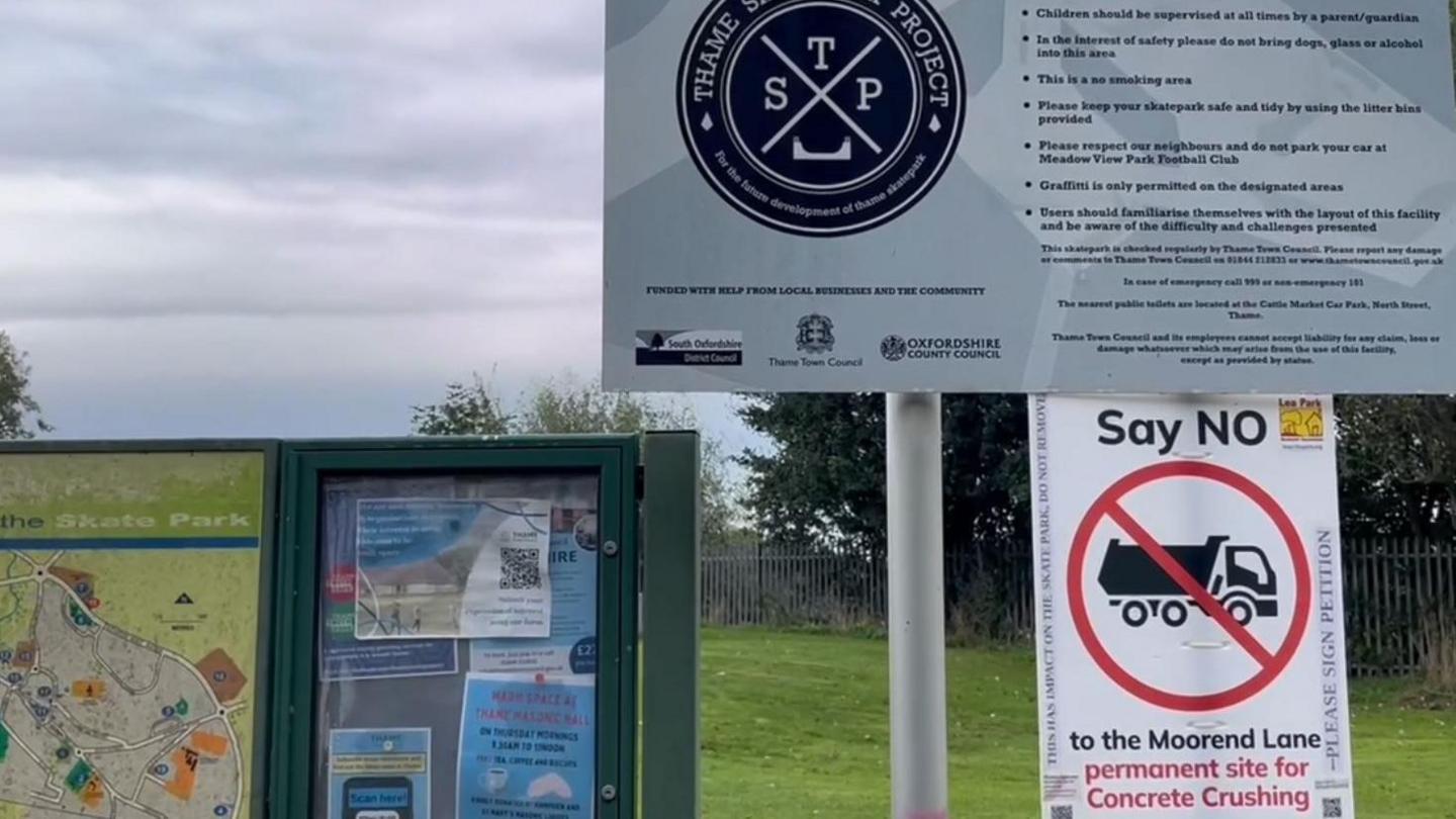 A placard reading "Say NO to the Moorend Lane permanent site for Concrete Crushing" glued to the bottom of the sign for Thame Skate Park. They are next to a community notice board with some posters. It's an overcast day.