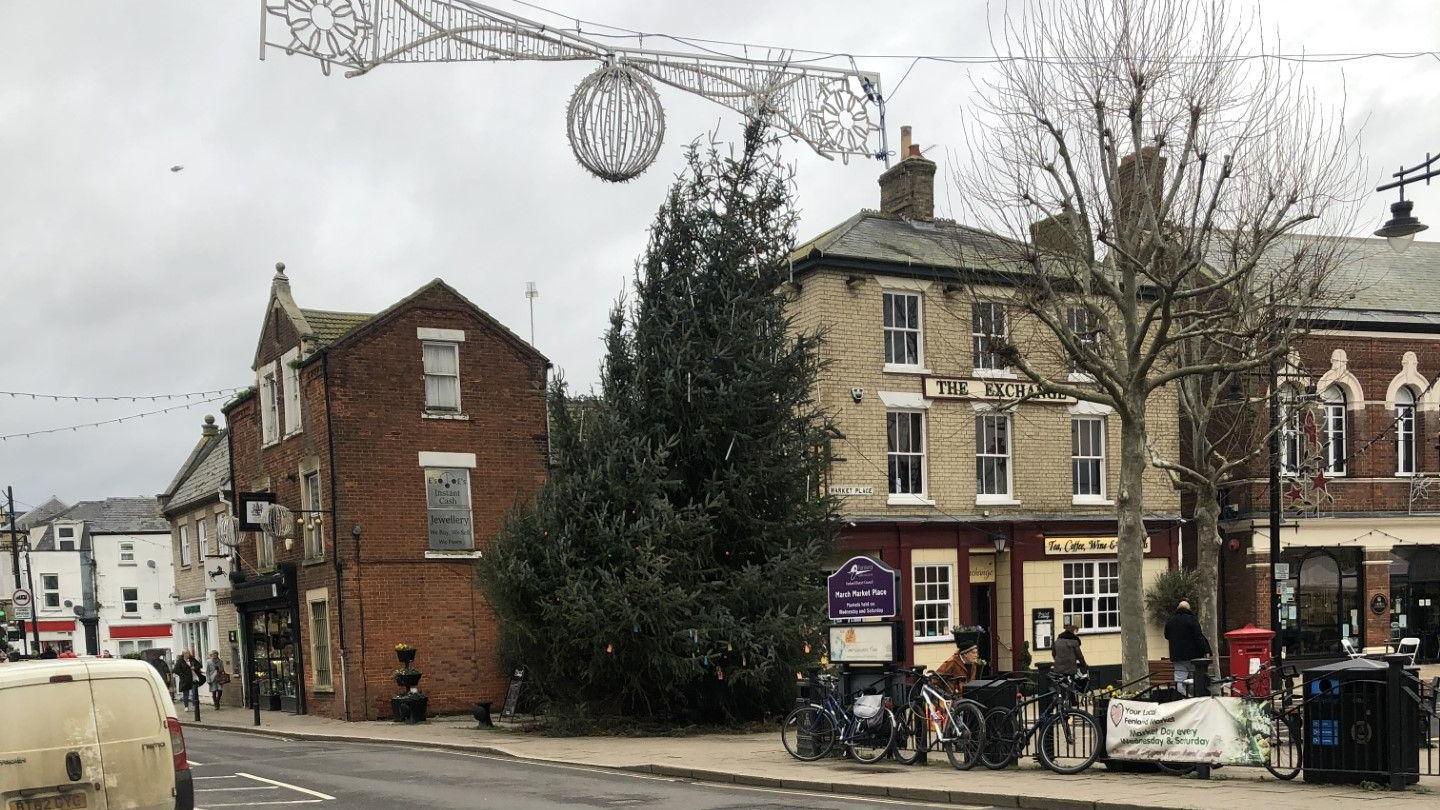 The market square in March, Cambridgeshire, various shops, cafes, bars behind a 9m (30ft) Christmas tree standing on a pavement, near the road with a pronounced lean to the right.