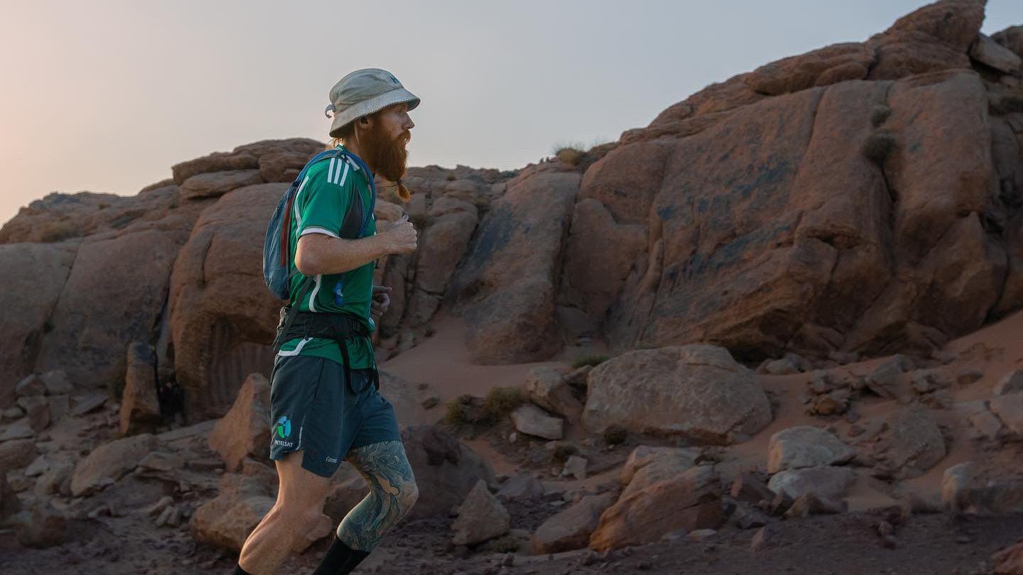 A man with a long red beard runs against a backdrop of red rocks in a bucket hat, green top and grey shorts.