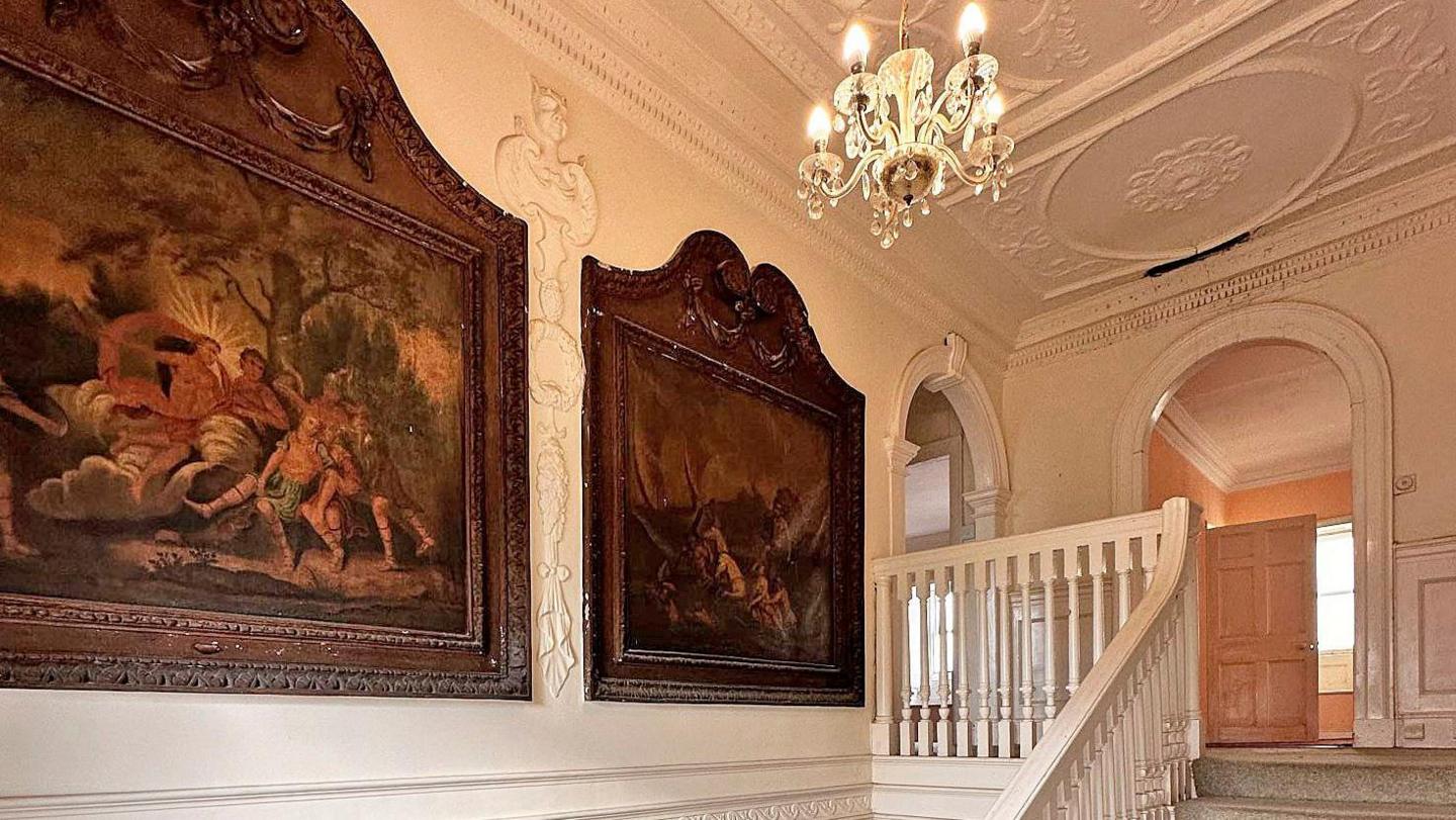 A view of an elegant white staircase leading up to a landing with white mouldings and large paintings set in decorative wooden frames.