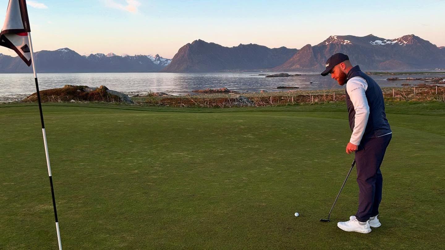 Isaac Rowlands is looking towards the hole and flag on the green of a golf course, it's about 10ft away. Behind him is open water with rocky mountains on the horizon.