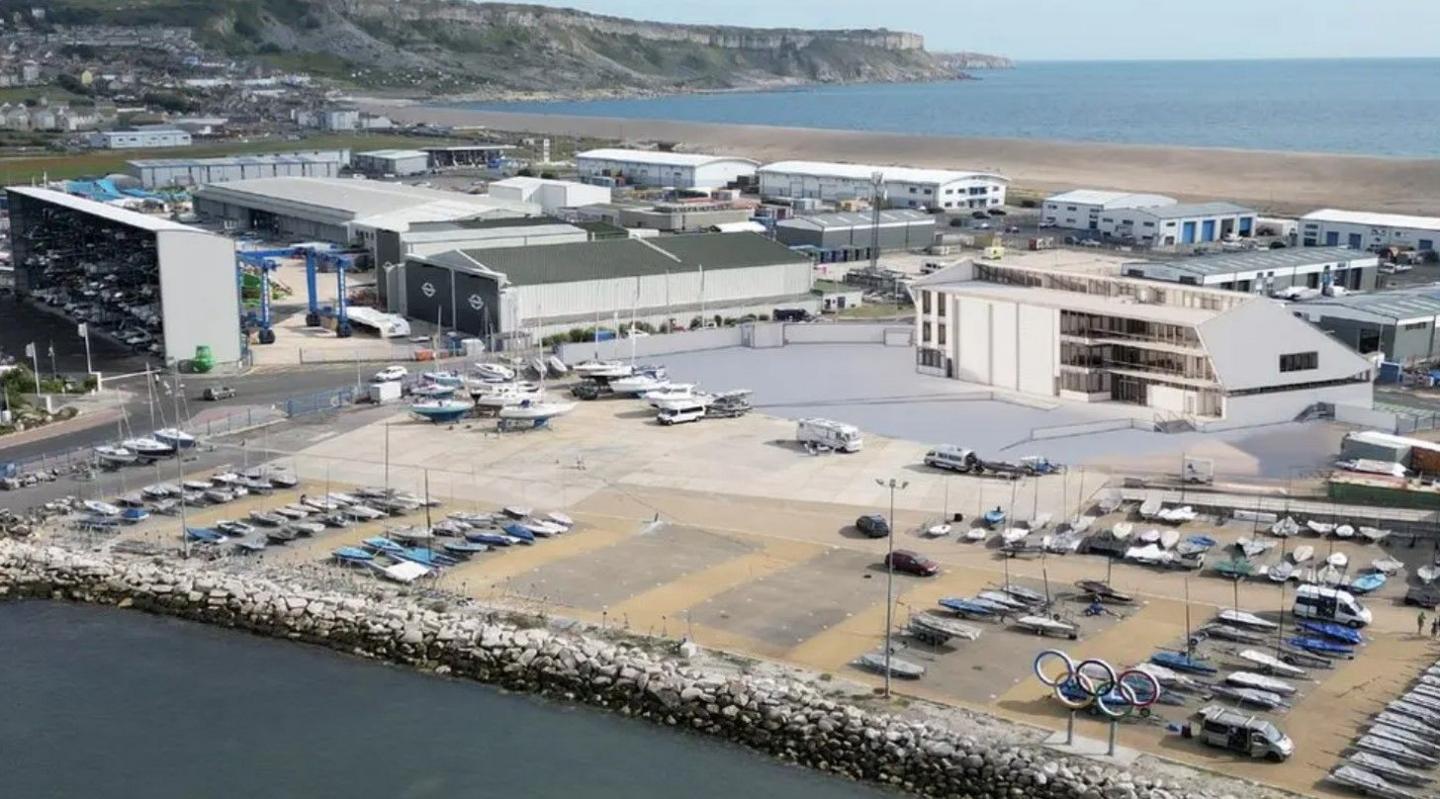 Aerial view of a state-of-the-art sailing training base - a white building with balconies to the front surrounded by a boat yard with cliffs, a a beach and the sea stretching behind