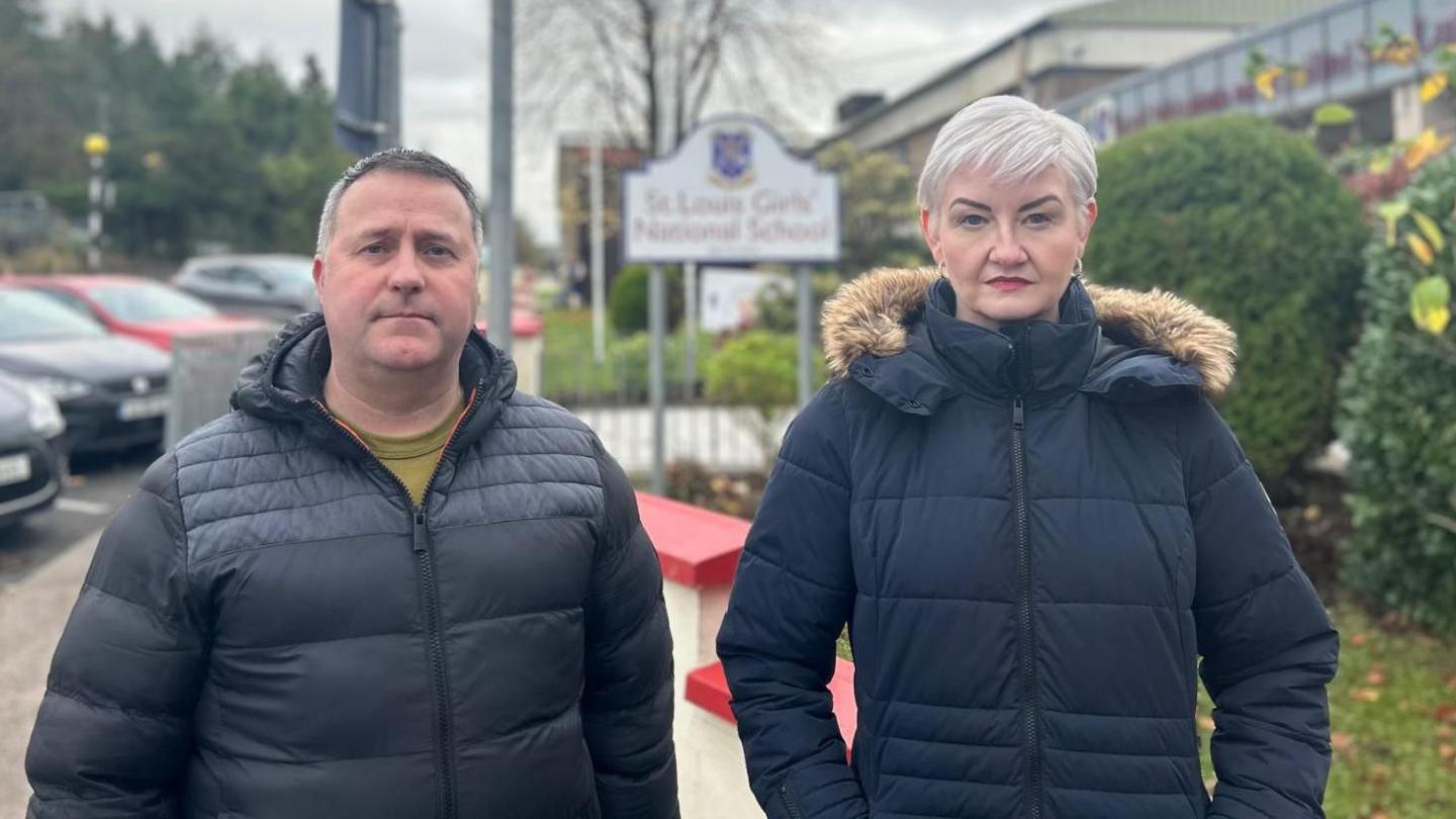 Sean and Julieanne Lynch stand in front of a low level cream wall which has it's top painted bright red. Behind them is an out of focus sign for St Louis Girl's National School, where their daughter was last seen alive. Julieanne is wearing a black duffle coat with her hands in her pockets. She has short blonde hair and red lipstick. Sean, who has short dark hair, is stood to her left in a black padded coat. Both are looking at the camera. 