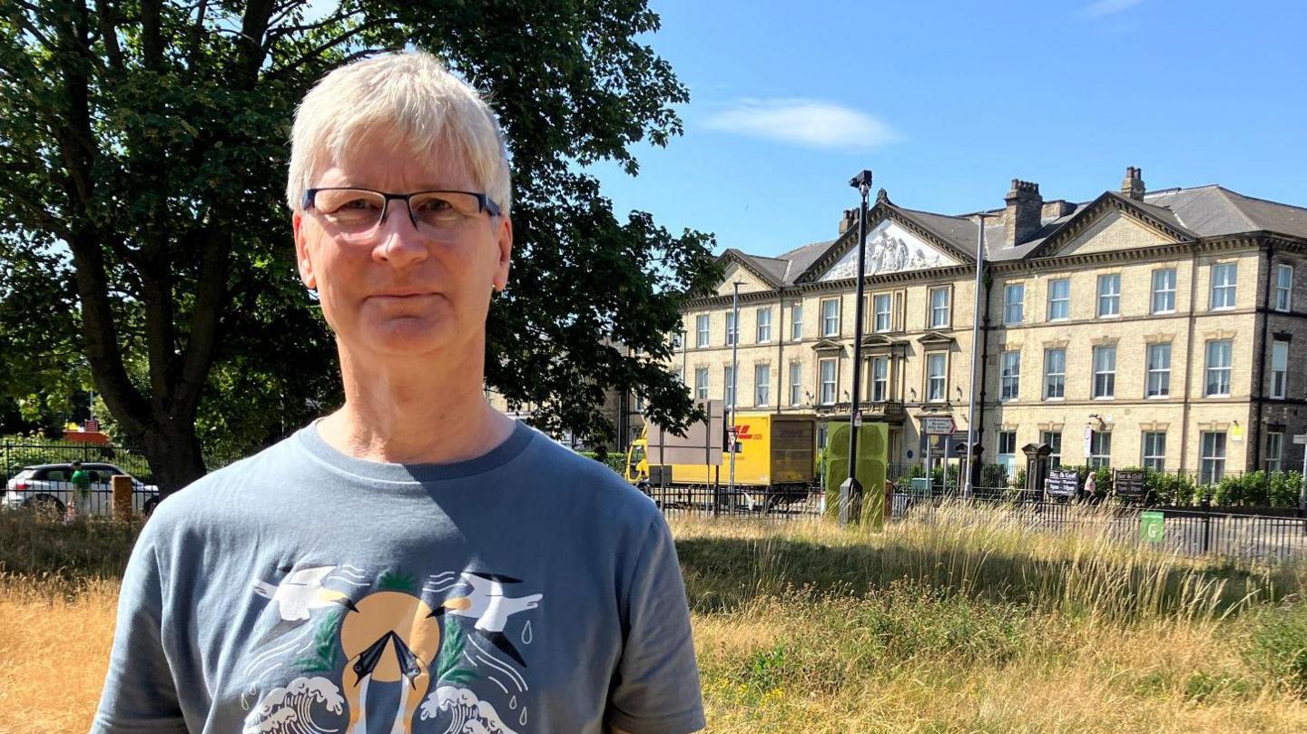 A man with white hair standing in a wildflower meadow. He is wearing glasses and a blue T-shirt with birds on it. Behind him stands a large, three-storey, Victorian-style building in yellow brick.
