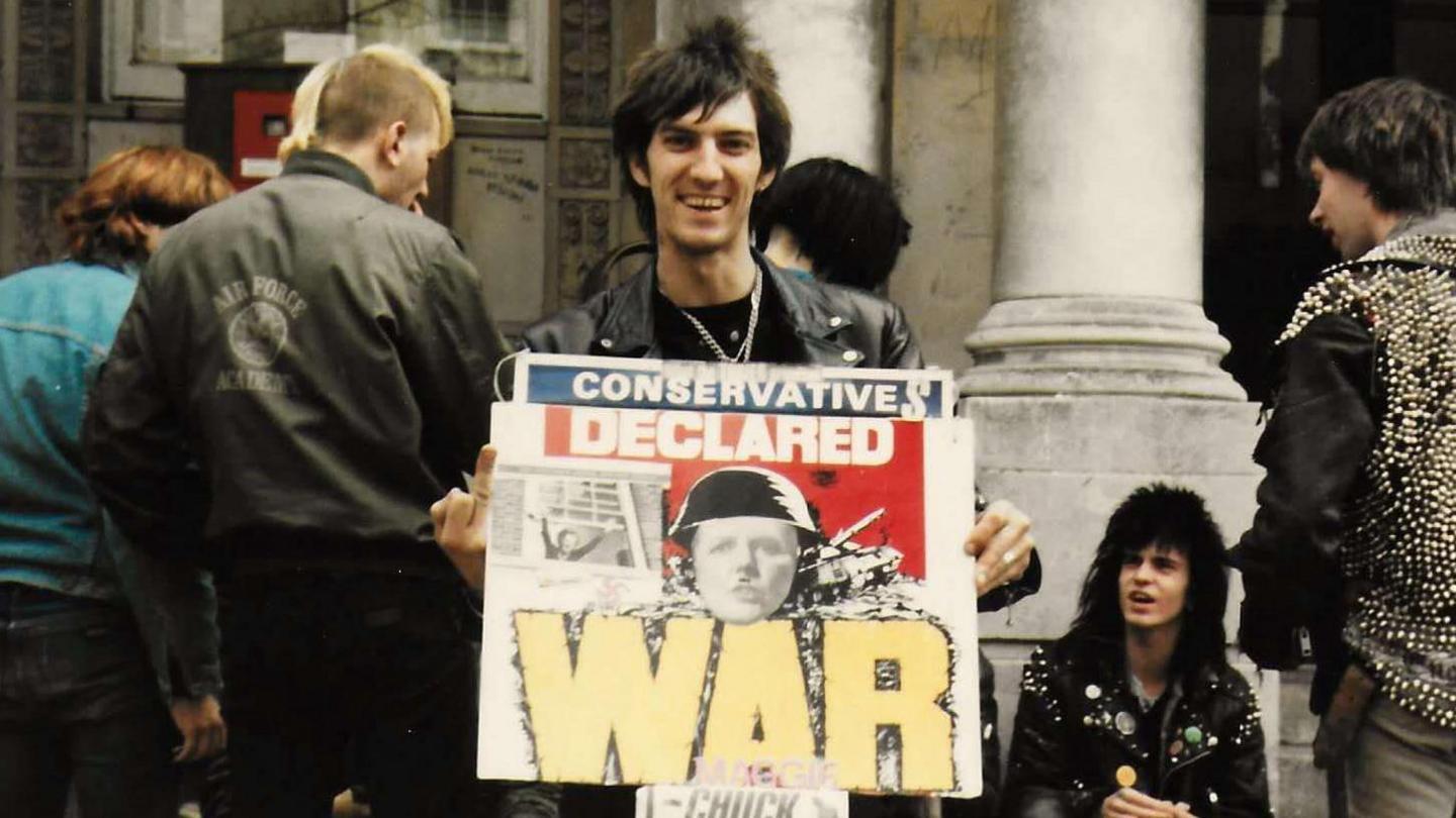 A man with long black hair wearing a leather jacket holding up a protest placard that says WAR on it