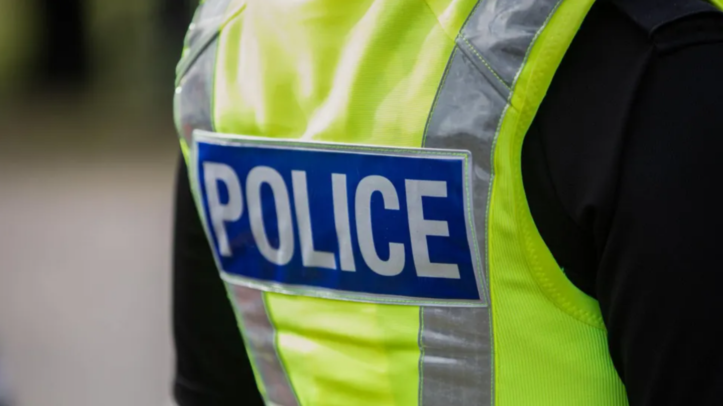 The back of a police officer. They are wearing black long sleeves, with a yellow hi-vis jacket with grey fluorescent stripes. There is a blue logo on the back, reading "Police"