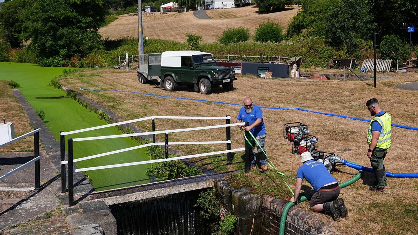 Three people, one in a high vis vest, direct a hose into the Hatton lock stretch of the Grand Union Canal in Warwickshire amid bright green canal water and brown, parched grass on its banks, with cars, houses and trees in the background on a sunny Wednesday afternoon.