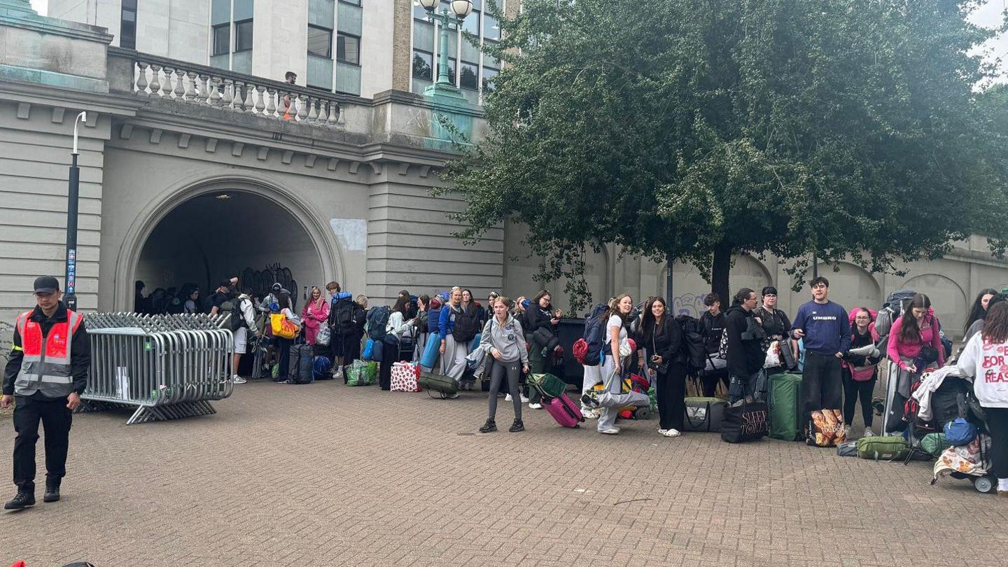 A long queue of young people with camping gear and backpacks waits outside a building near an arched tunnel.