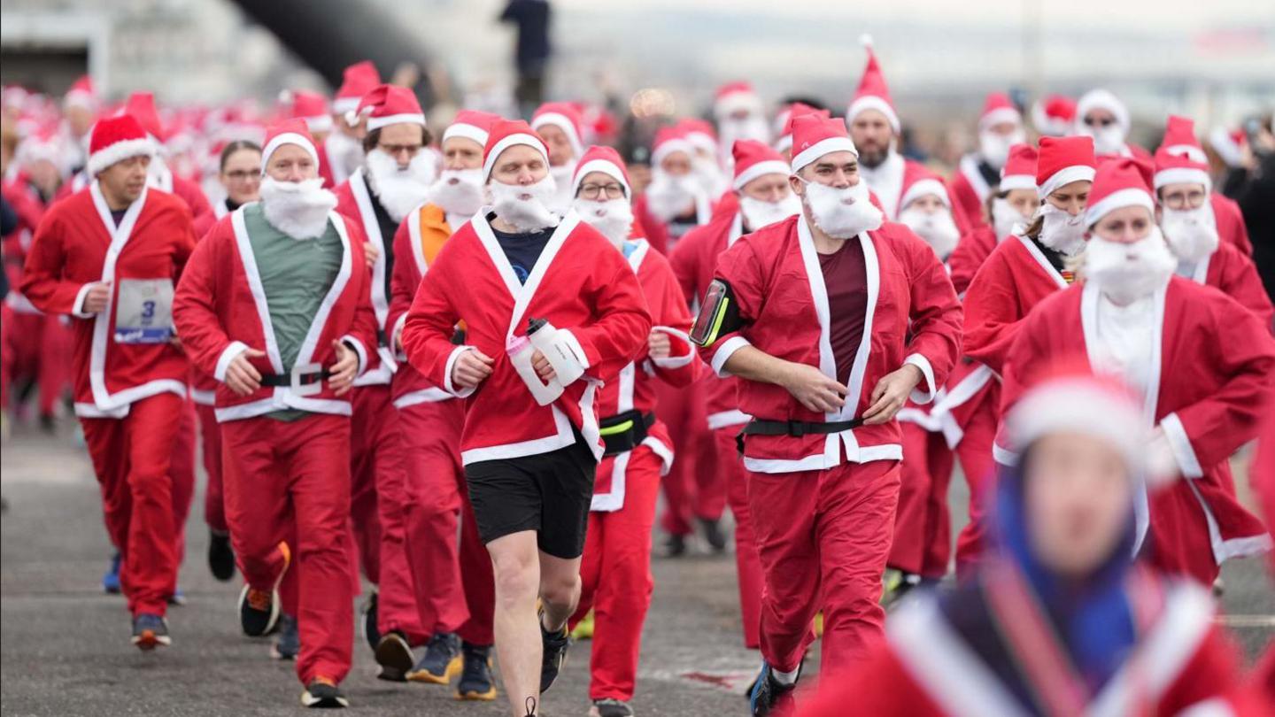 Dozens of people running while dressed as Santa Claus. All are wearing red and white outfits, a white beard and a Christmas hat.