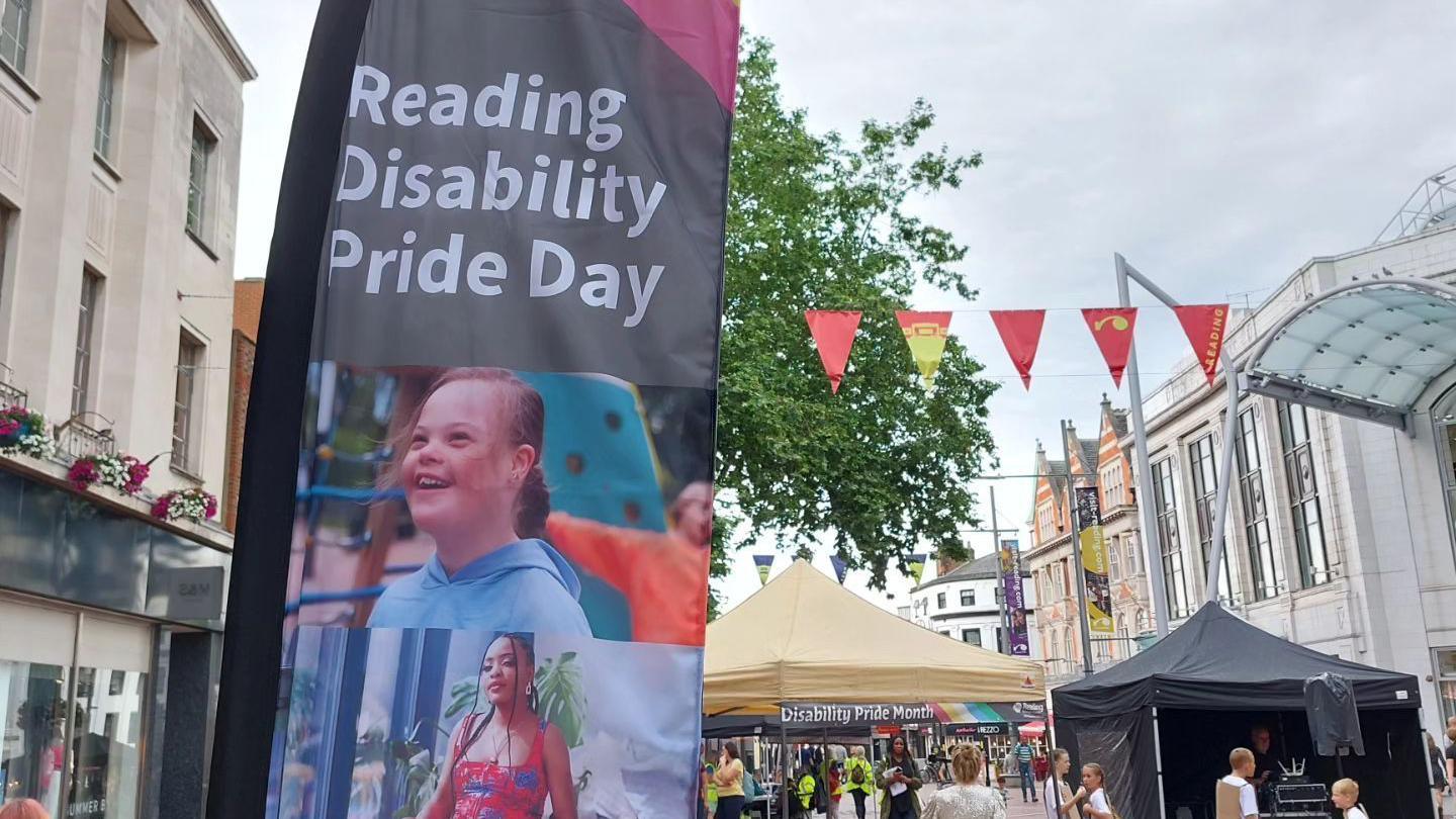 A sign reading "Reading Disability Pride Day". There is a girl smiling on the sign. To the left, there is red and yellow bunting. In the background is a gazebo with a light yellow top.