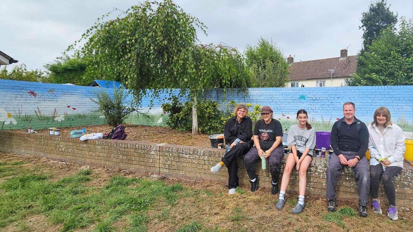 Local artists sit on a wall in front of the wildflower haven mural which is light blue with grass and insects