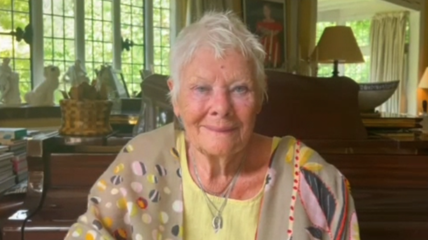A slightly grainy image of Dame Judi Dench, smiling at the camera. She is wearing a patterned jacket and pale yellow shirt. She is sitting inside a cluttered room, with an upright piano behind her, lamps, books and ornaments on the windowsill.