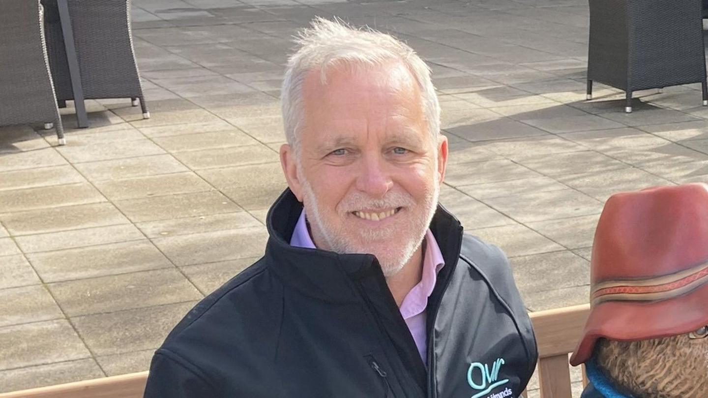 A man with grey hair and a short grey beard wearing a navy jacket over a pink shirt