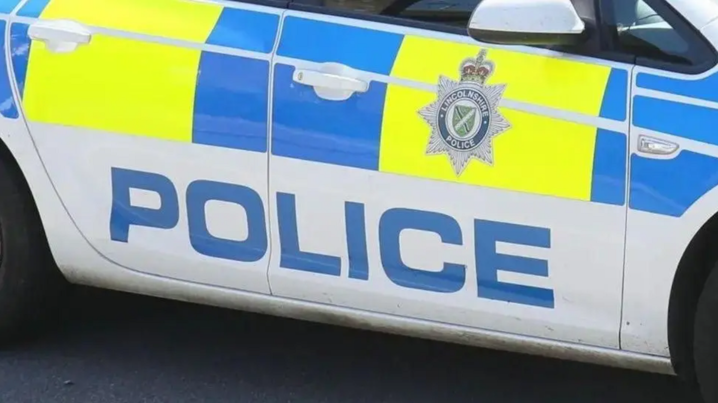 A side view of a marked Lincolnshire Police car. It is white with yellow and blue blocks and has the force's badge on the door.