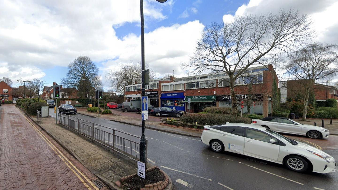 A street view image showing a row of shops in the background and a main road in the foreground with two white cars to the right of the frame.