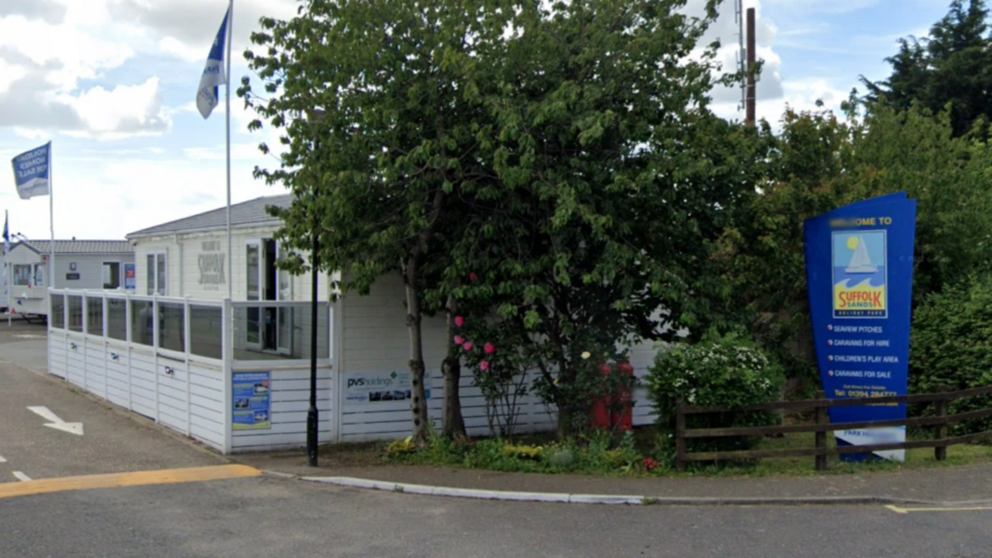 The entrance to a holiday park with a blue sign reading "Welcome to Suffolk Sands", and flags running down the driveway.
