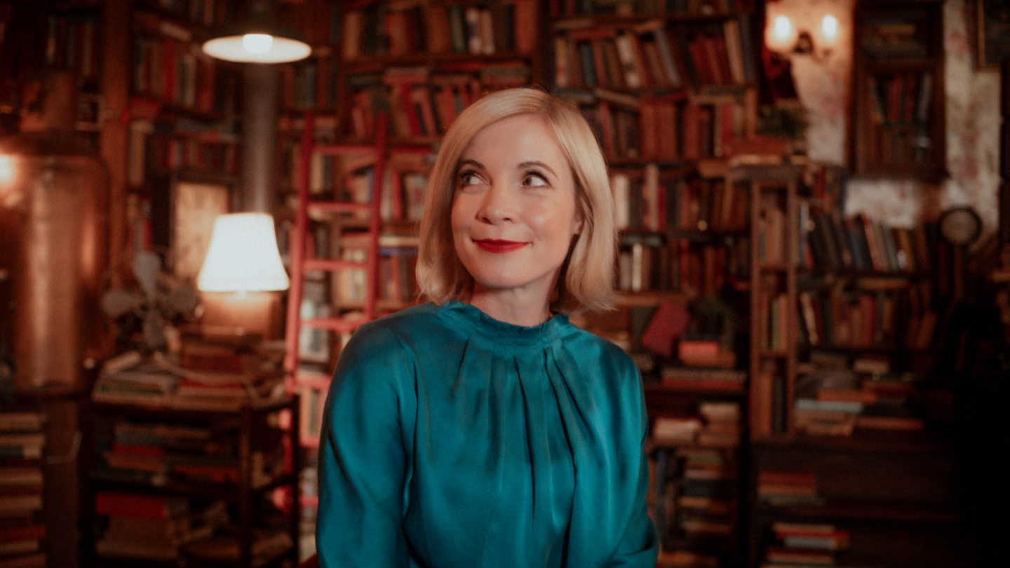 Lucy Worsley has shoulder-length blonde hair and wearing a turquoise outfit. She is in a room filled with old books.