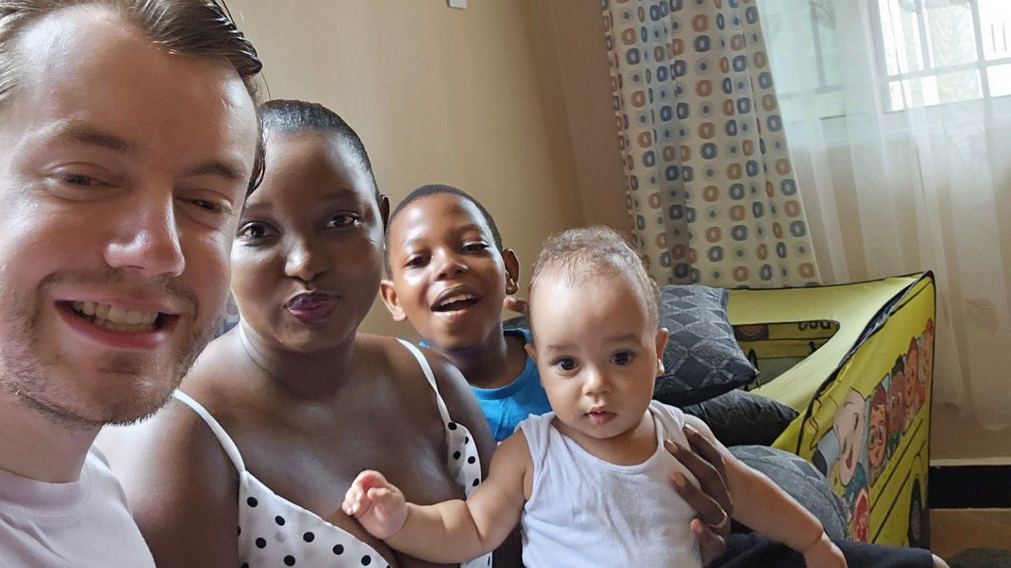A family of four smiles for a photo inside a living room. A man, a women, and their two young children aged 11 and two. They seem happy together.
