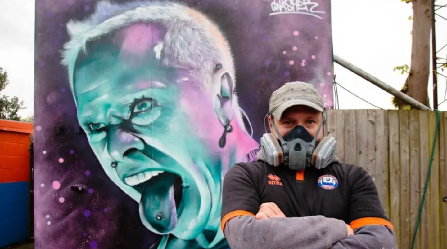 David Nash, wearing a face mask, standing in front of his Keith Flint mural - a purple and blue painting showing Keith shouting with his pierced tongue sticking out.