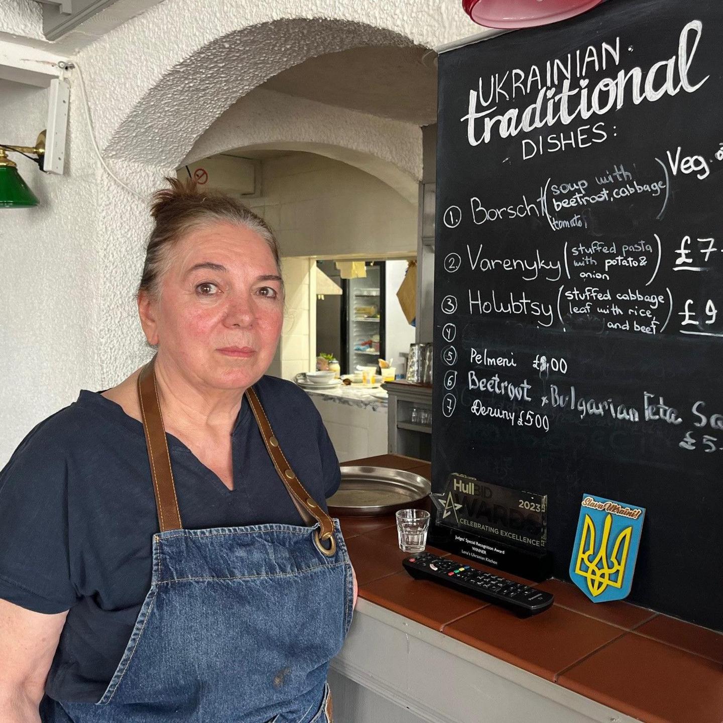 A concerned-looking Lena Sutherland, the owner of Lena's Ukrainian Kitchen. She is wearing a denim apron in front of a menu board listing a range of traditional Ukrainian dishes 