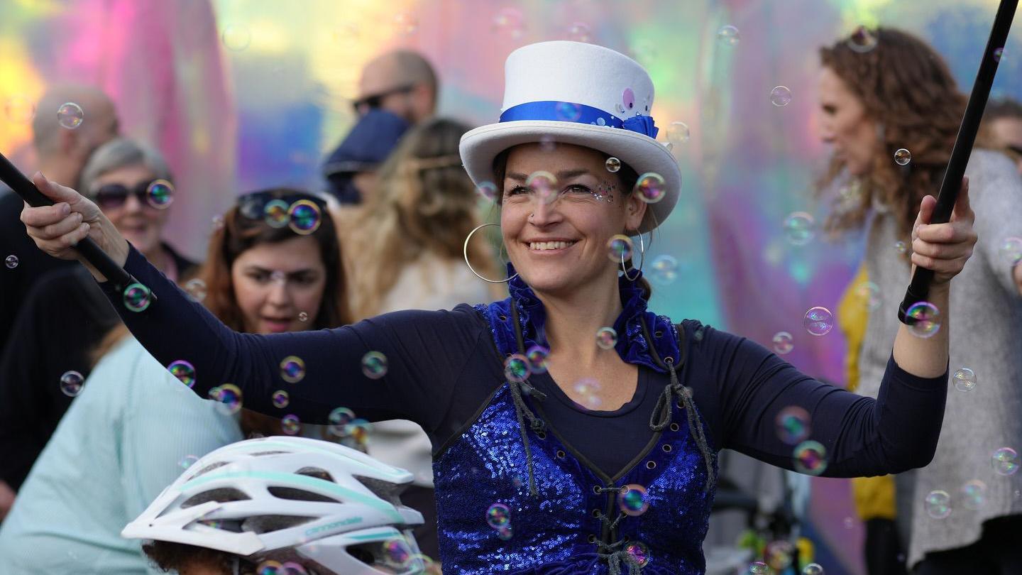 A woman in a top hat and sparkling blue outfit walking with canes in each hand which she is holding aloft. She is smiling as she walks through crowds of people and bubbles in the air.