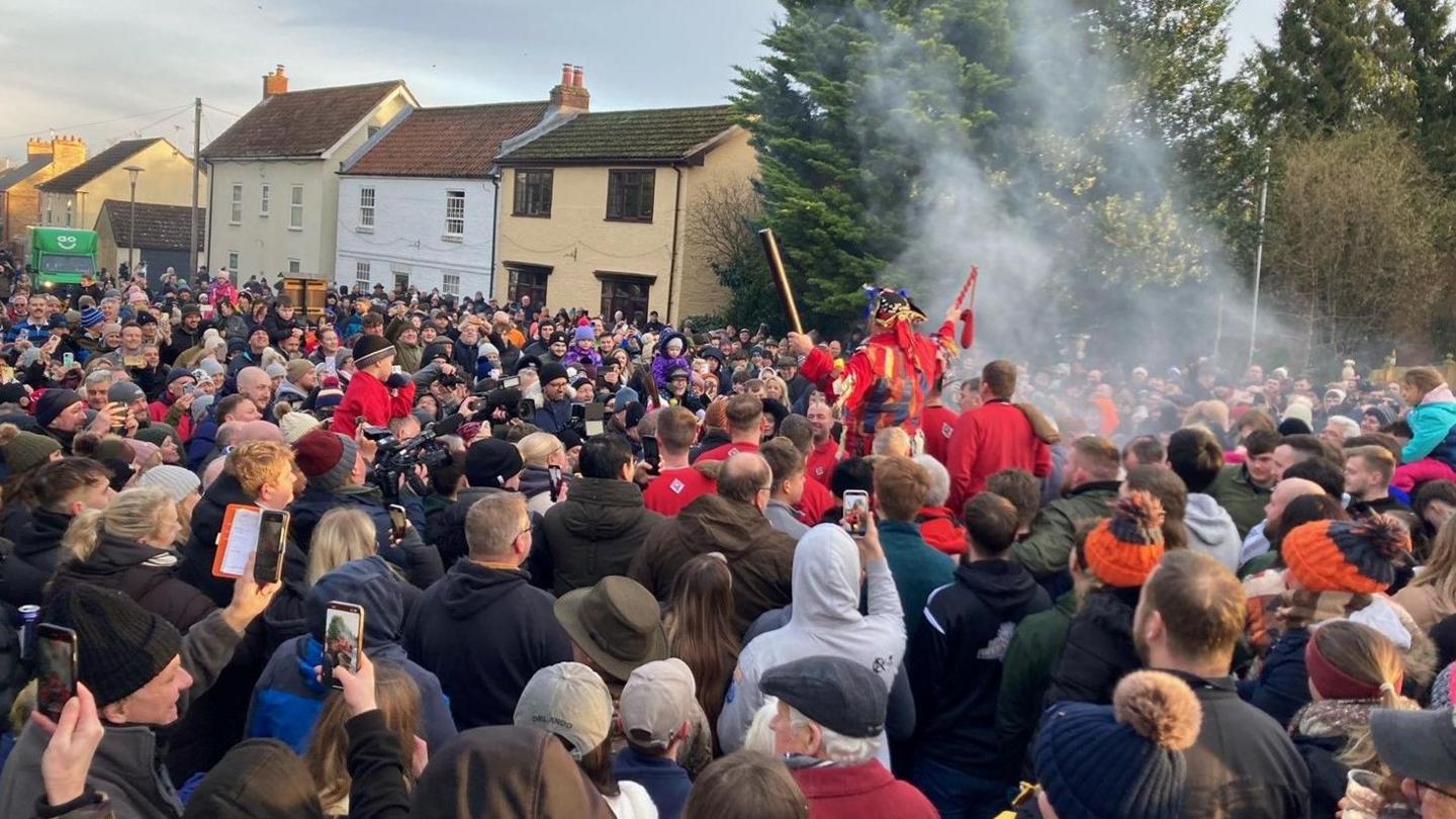 A large crowd of people prepare to take part in a medieval rugby-style game in the centre of a village. They are surrounding the fool, who is dressed in red and holding a leather tube (the ball) aloft. Some people are filming the spectacle on their phones. Houses and trees can be seen in the background.