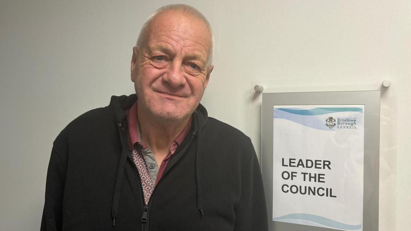 Milan Radulovic wearing a black hoodie and red collared shirt in front of a sign which says leader of the council