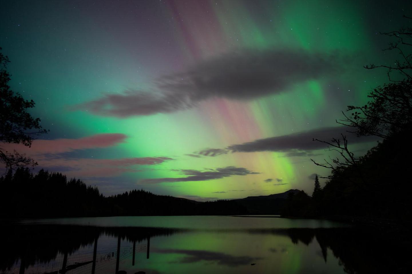 Green and pink glow of the aurora behind clouds and reflected in the waters of a loch.