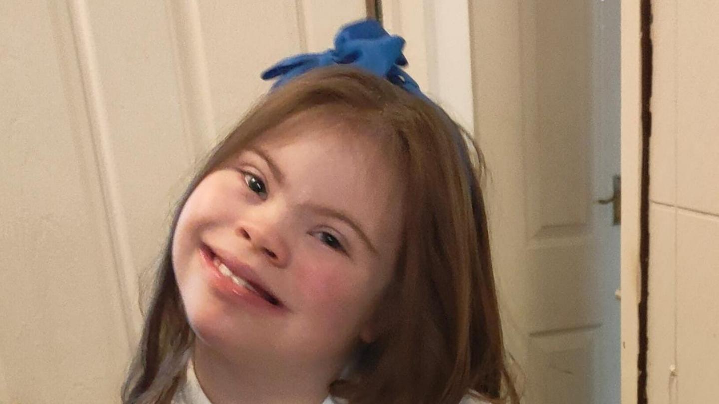 A young girl with long brown hair styled with a blue bow, smiles