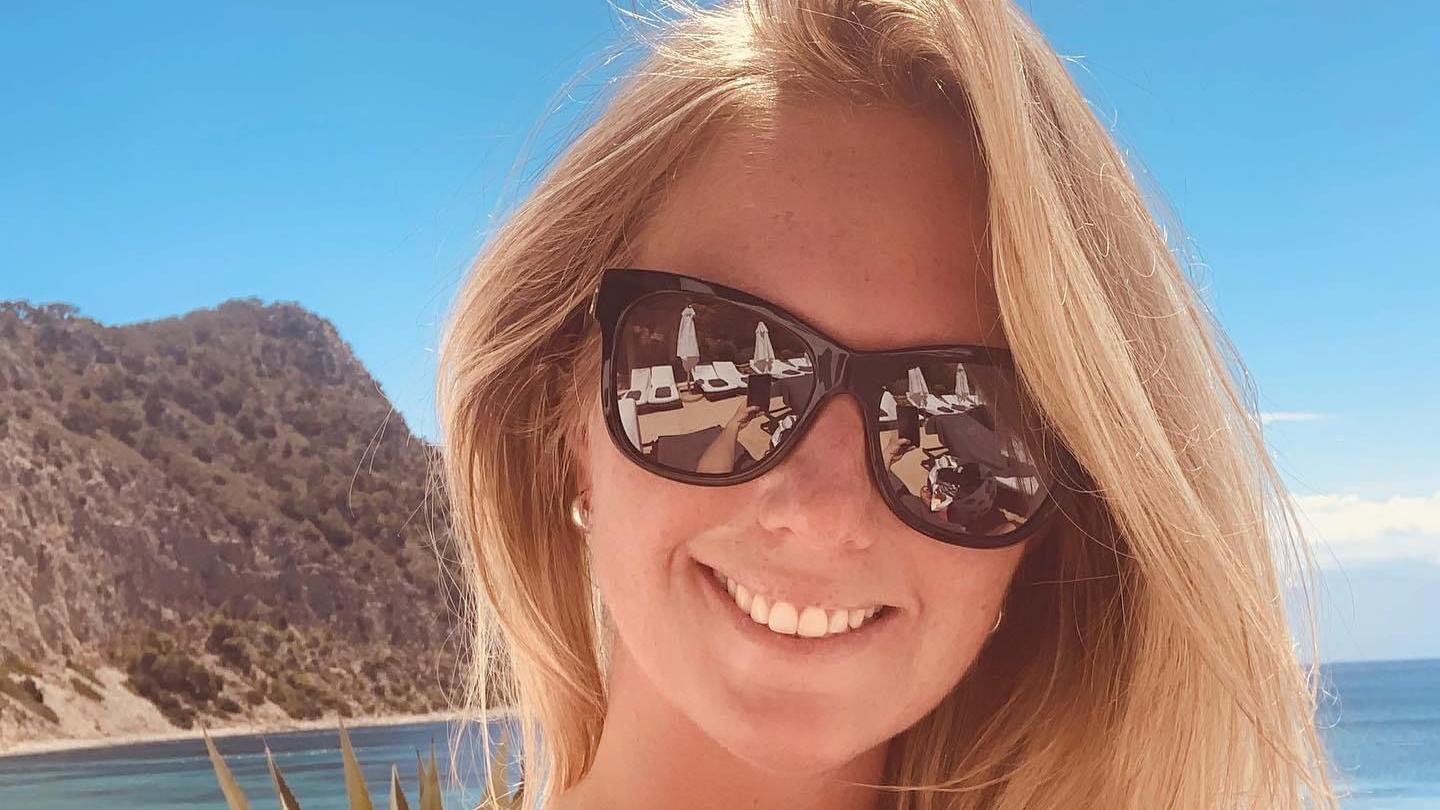 Gemma Willis, who is smiling and wearing black sunglasses. She has long blonde hair and is on a beach. A large hill is over her shoulder, with the sea also behind her. It is a sunny day.