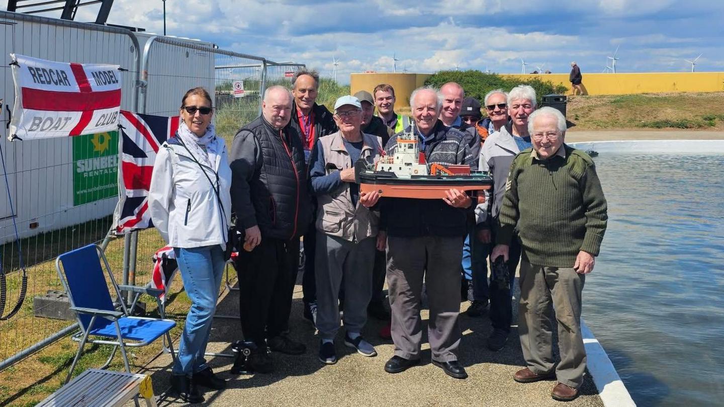 About a dozen members of Redcar Model Boat Club smiling next to the partially refilled lake. A member in the centre of the group is holding a brown and white model boat. There's a flag on the side of a portable building next to them, with the name of the club on it.