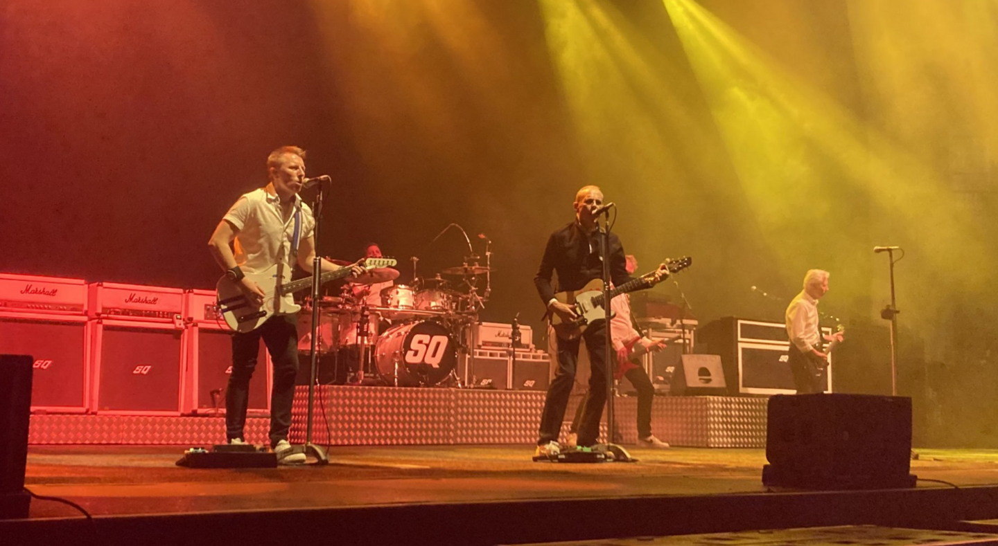 Images shows band mates singing into microphones and playing guitars. The stage has red and yellow lights shining on it