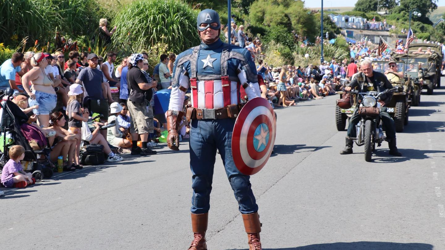 A man dressed in a Captain America outfit taking part in the carnival, with people to his right watching on.