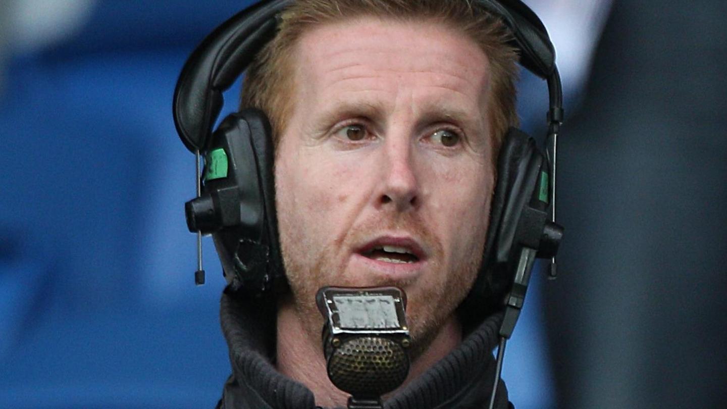 Iwan Roberts is in a football stadium and is using a commentator's microphone and is wearing headphones. He is looking to his left. He has short spiky, ginger hair and ginger face stubble.