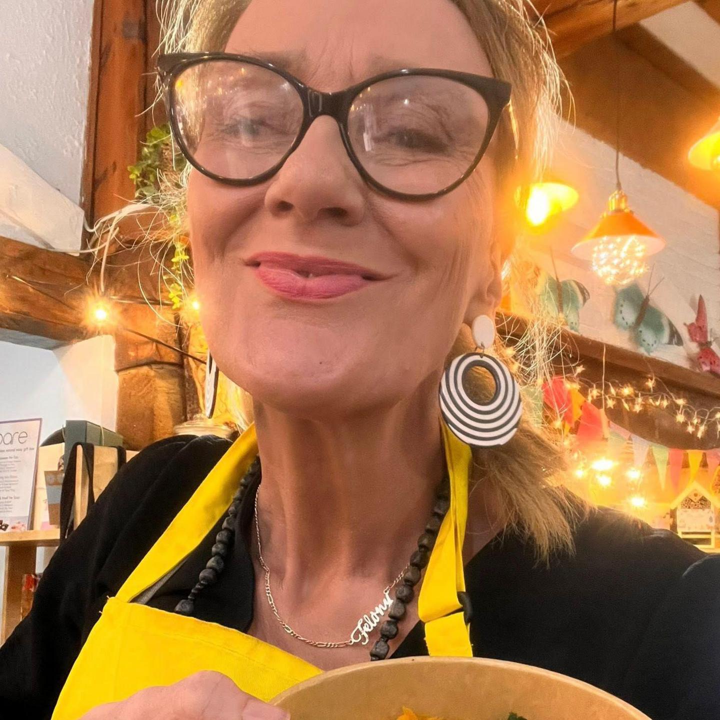 Image of Mel. She has a yellow apron on holding a bowl of food to the camera.