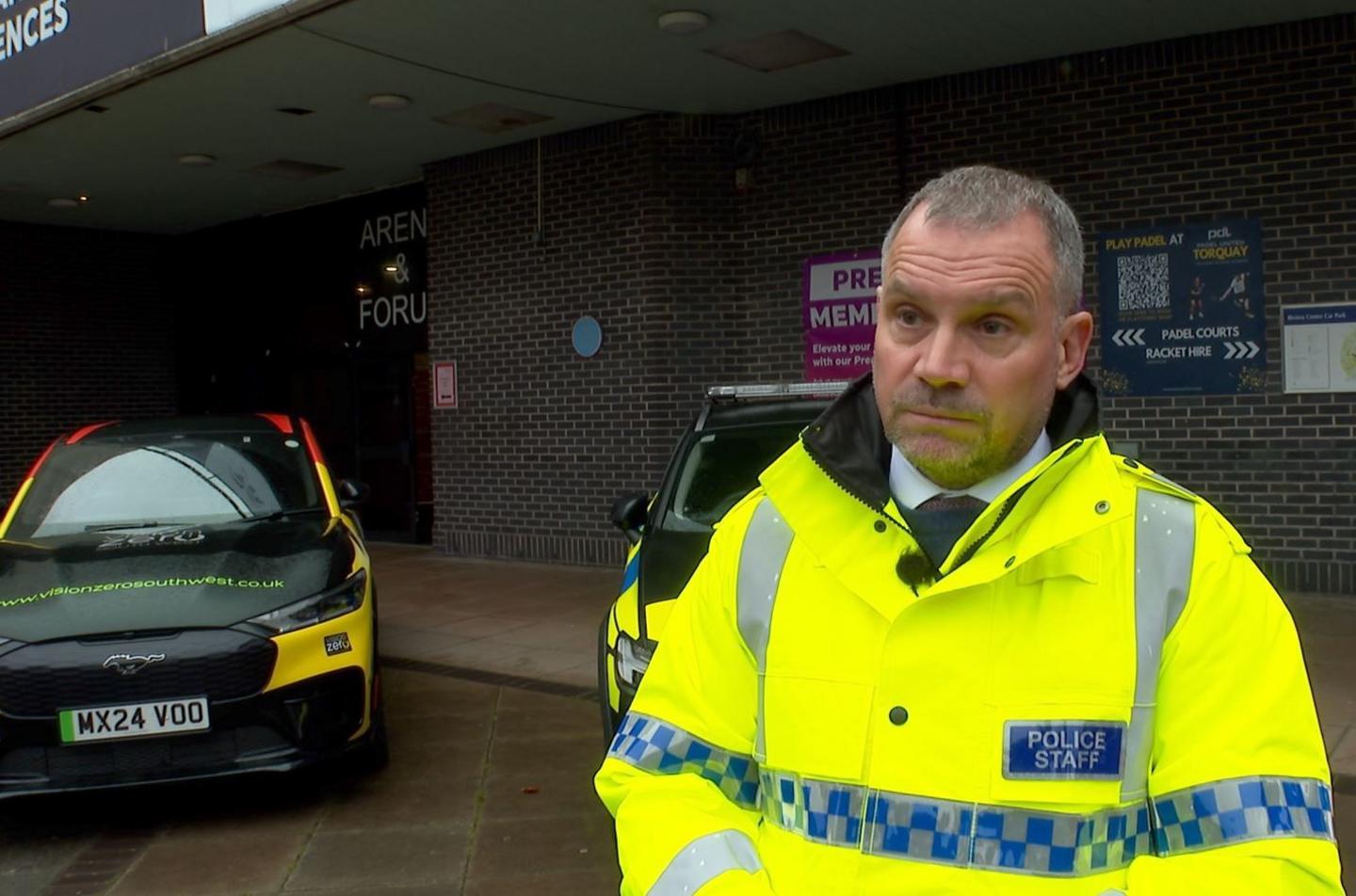 Adrian Leisk wearing a high-visibility yellow jacket labelled "Police Staff" and standing in front of a building. Behind him are two vehicles, one of which is branded for "Vision Zero South West" with a distinctive yellow, black and white design. Another vehicle is partially visible to the side.