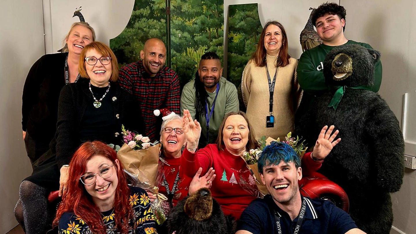 A group of shop workers, wearing festive outfits, smiling towards the camera. There is a black stuffed bear on the right hand side of the photo.