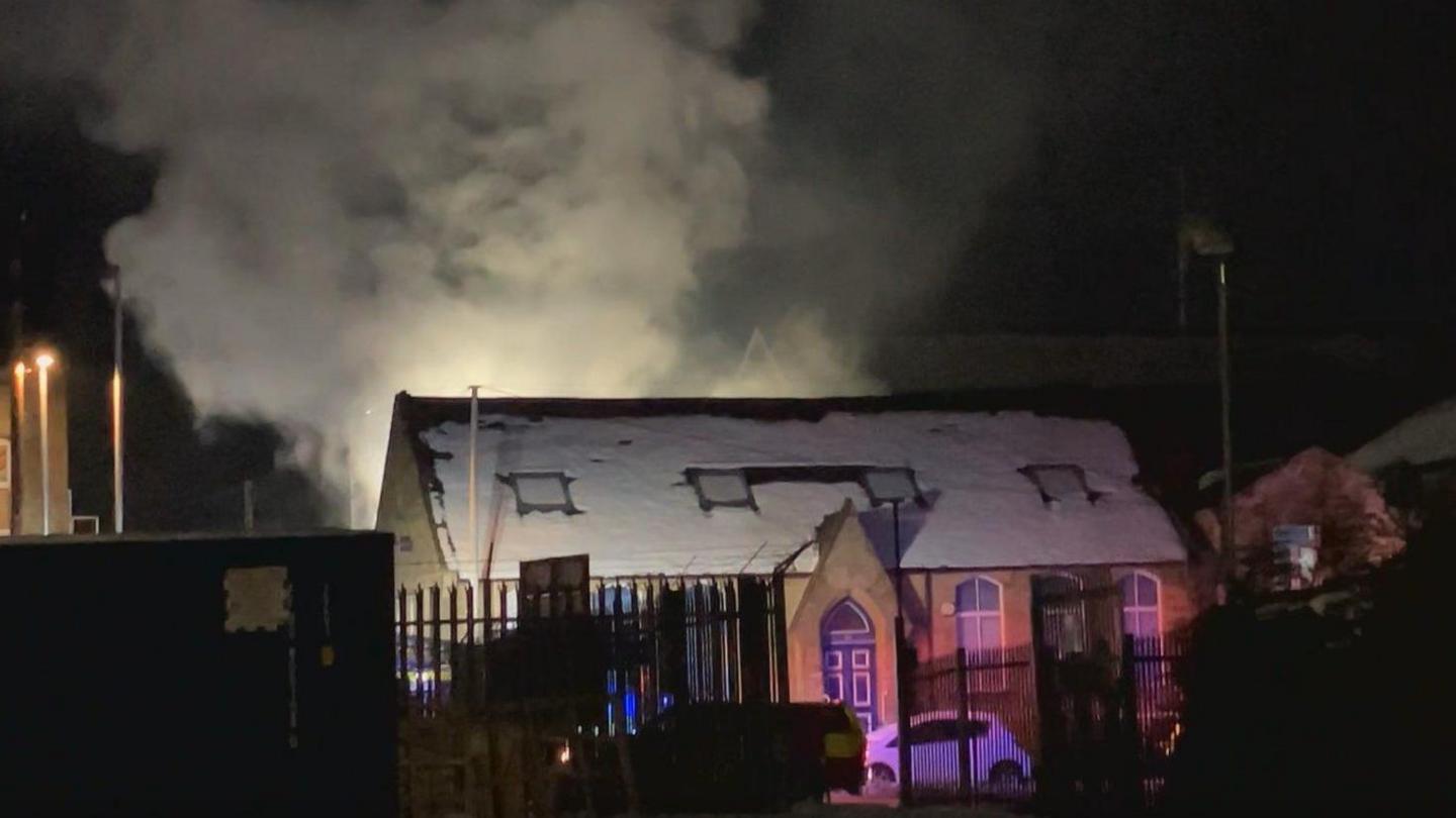 A large plume of smoke rises from a building with snow on its roof.