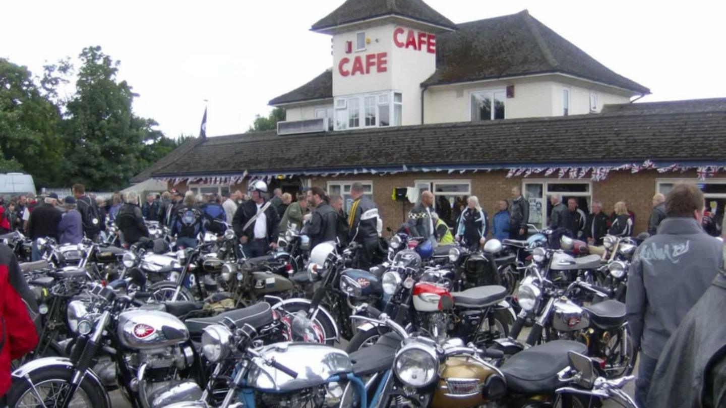A cream-painted two-storey cafe. The lower floor has white-framed windows and a pitched roof. The upper floor is much smaller and has an entrance with a small tower which has the word CAFE in red lettering. There is a large group of motorbikes in front of the building, covering all the space in the car park. Some bikers can be seen among the vehicles.