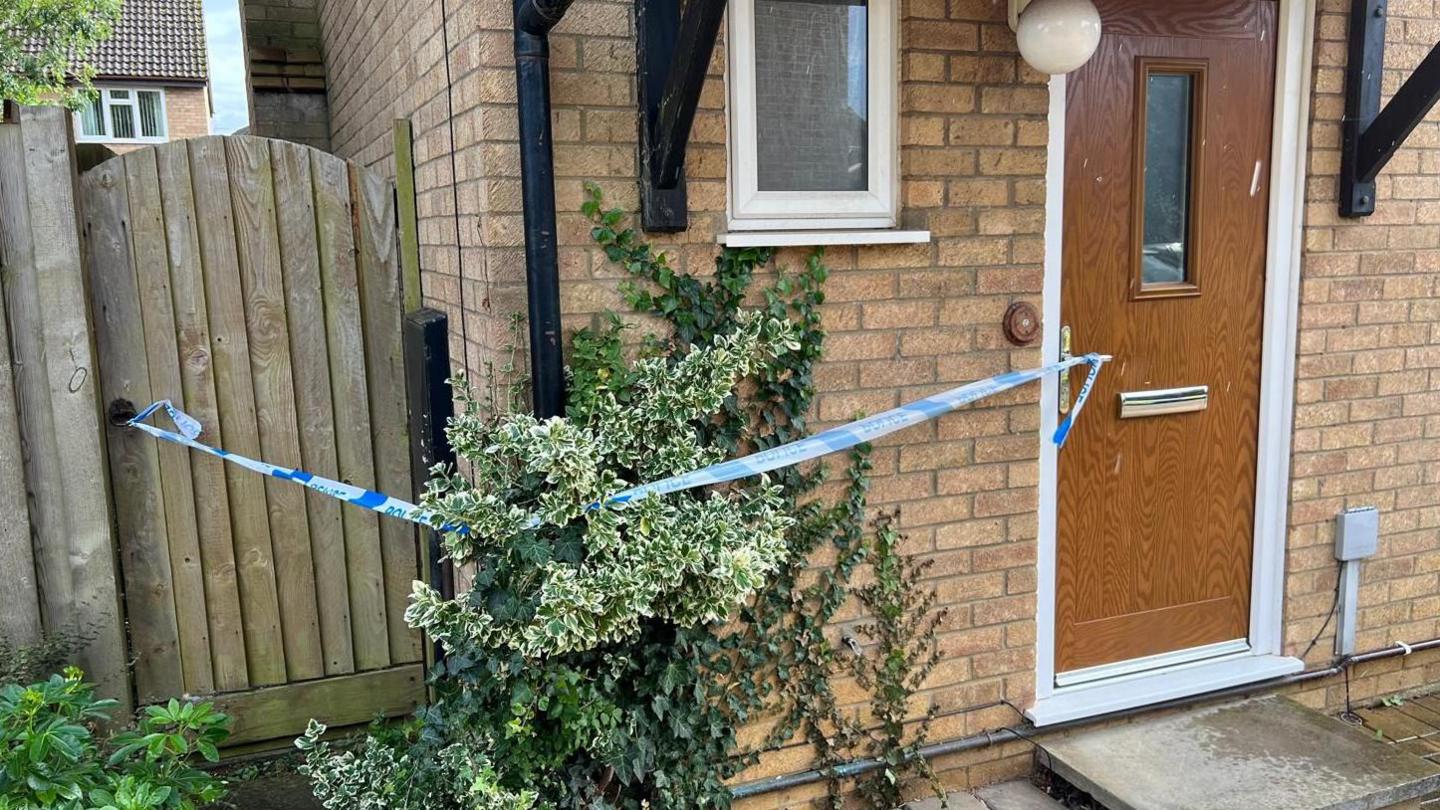 A close-up of a brown wooden front door with blue and white police tape running from the front door handle to the garden gate handle to its left, John Amner Close, Ely