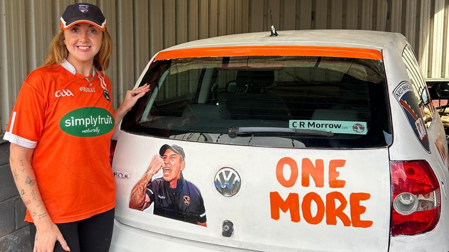 Gillian Kelly wearing an Armagh GAA top standing beside a white car that has been decorated with orange paint and a picture of the Armagh manager.