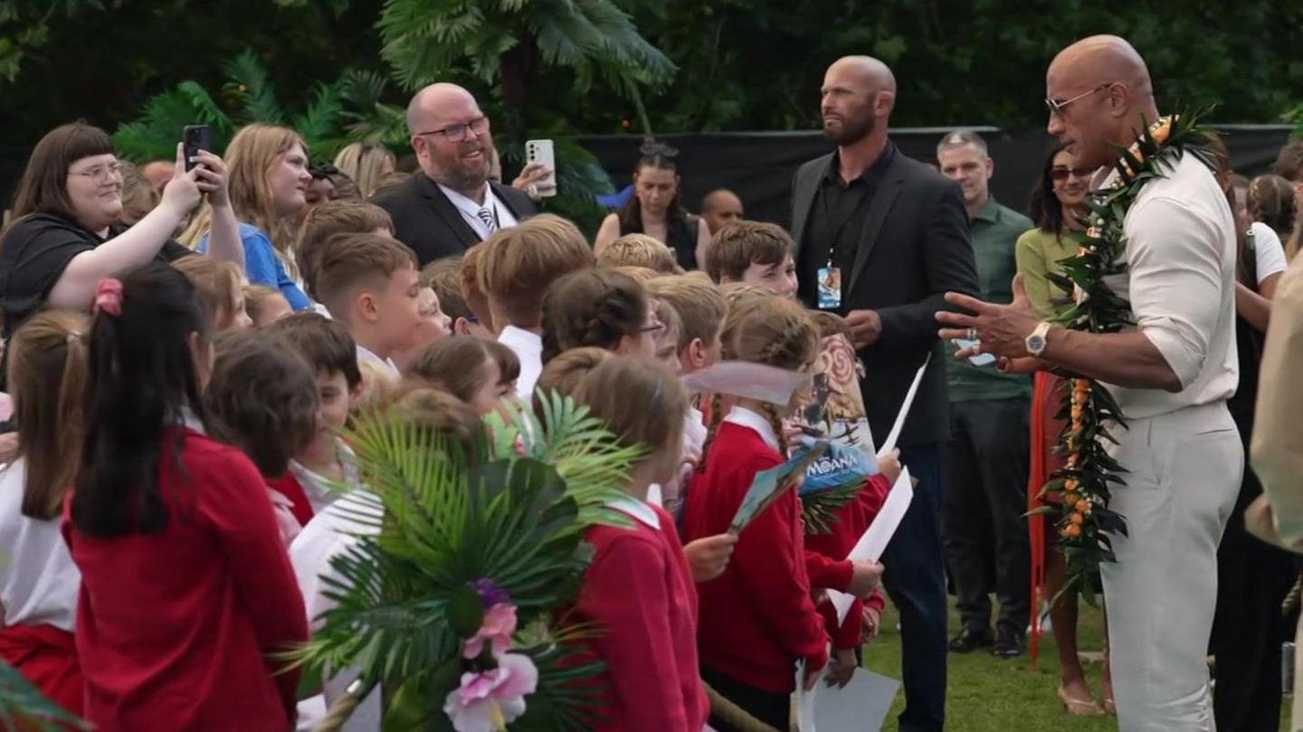 A radical  of schoolchildren wearing reddish  uniforms lasting  successful  beforehand   of The Rock – a antheral   wearing a pick  jumper and achromatic  trousers and sunglasses.