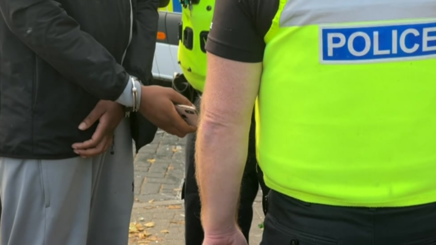 A police officer in a yellow high visibility vest talking to a person in grey tracksuit trousers