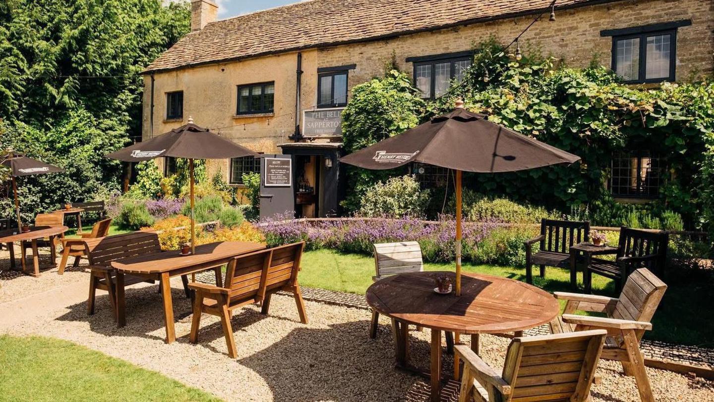 A photo showing a pub garden with a Cotswold stone building in the background. It shows a number of tables with umbrellas and chair around them