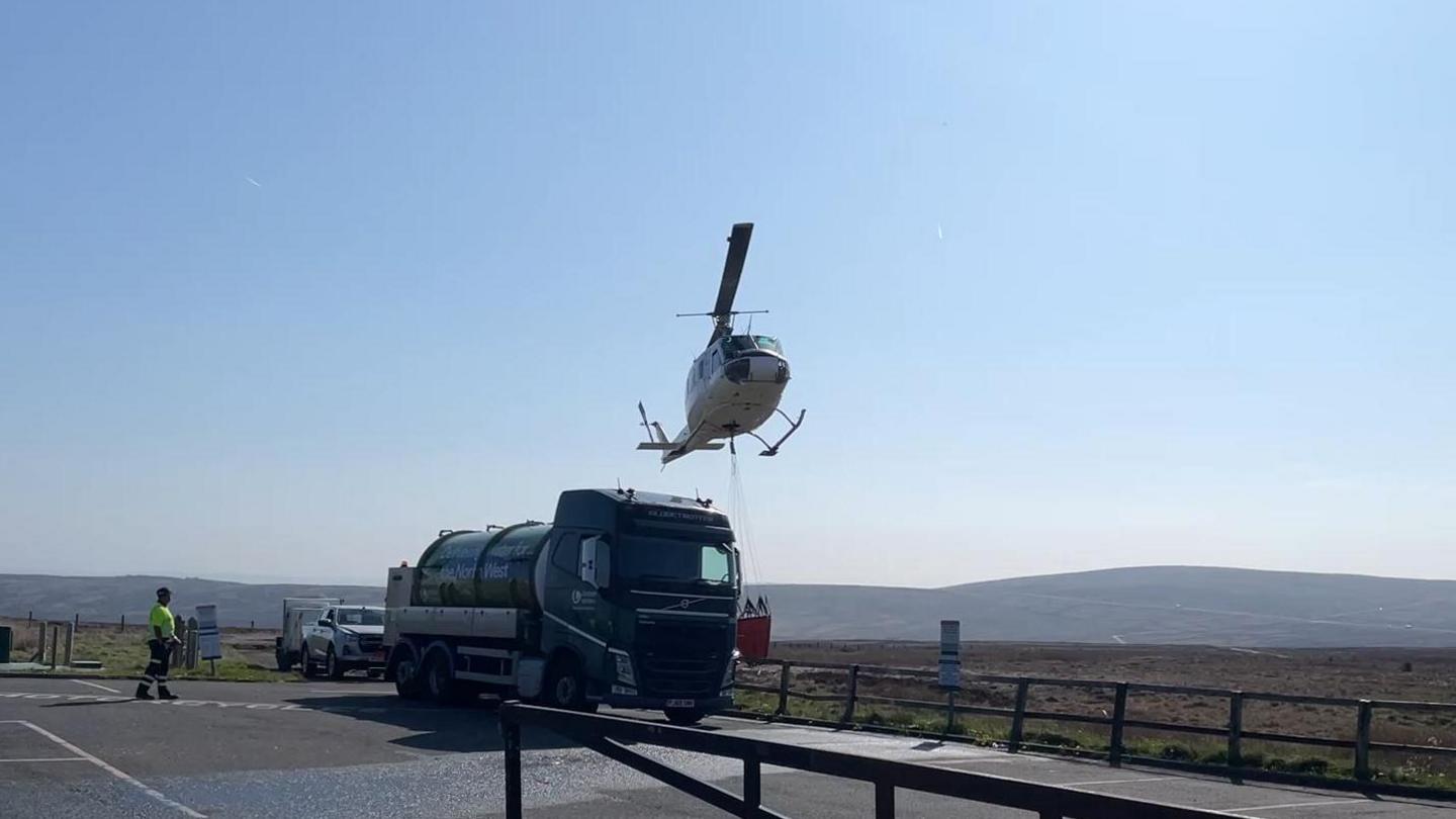 Helicopter hovering over a water bowser in a car park with moorland in the background