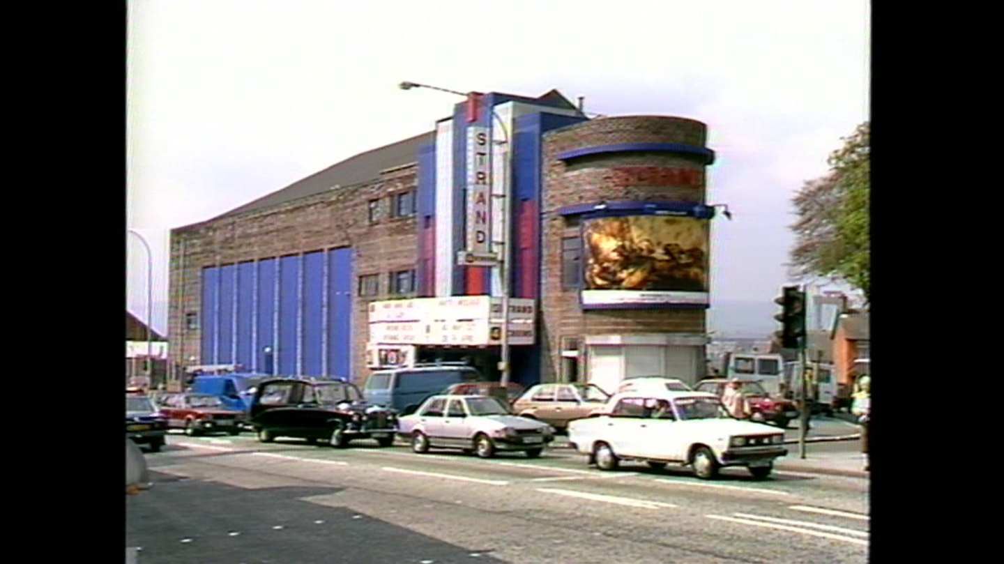 An archive image of the Strand Cinema situated on the corner of two perpendicular streets. It's signs are in blue, red and white. There are some classic cars on the road. 