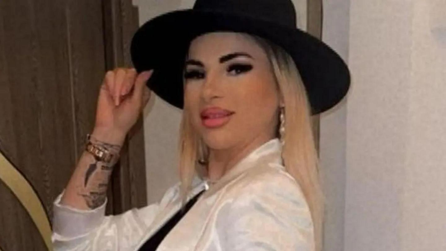 A woman, with blonde hair, wearing a black hat and white shirt. She is holding the hair with one hand. She has tattoos on this arm. She is smiling at the camera.