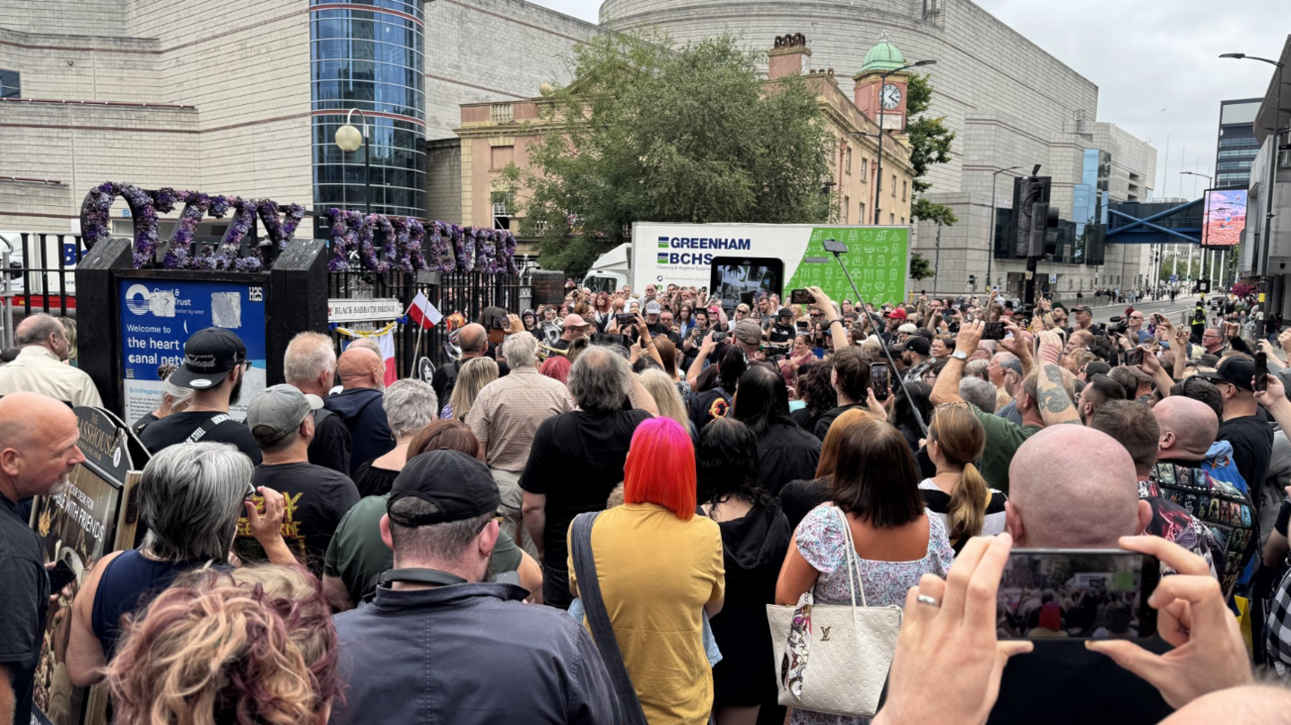 Hundreds of people crowded around the Black Sabbath bench in Birmingham. A purple floral arrangement reading 'Ozzy Forever' is visible above the bench.