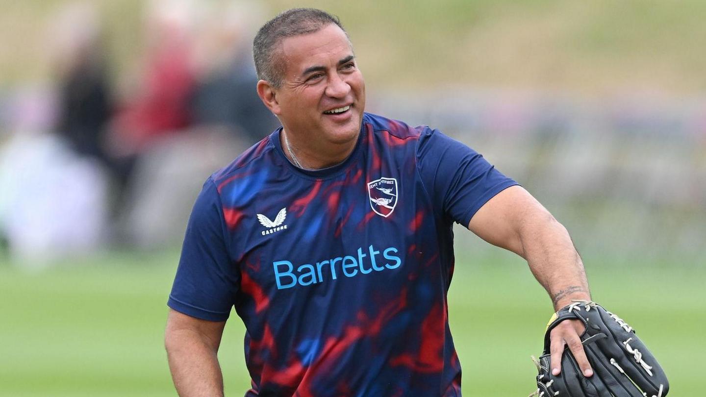 Adam Hollioake wearing a blue shirt, baseball glove and smiling